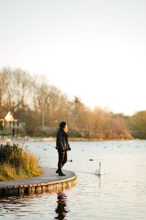 girl in sunset light stood by a lake wearing a beret and a faux shearling aviator jacket