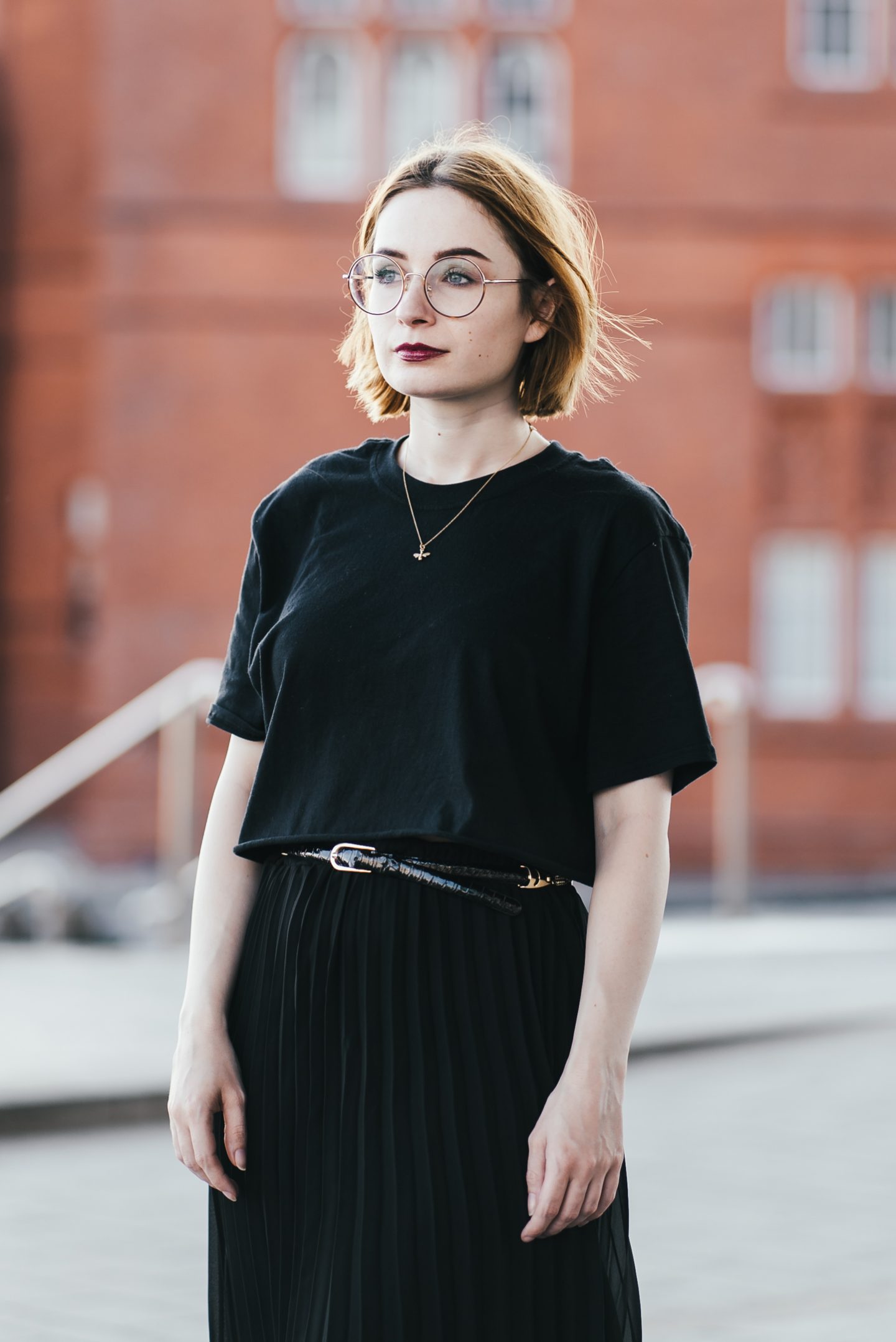 girl wearing a black t shirt, black pleated skirt and gold bee necklace with short ginger hair and round glasses