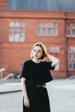 girl wearing a black t shirt, black pleated skirt and gold bee necklace with short ginger hair and round glasses