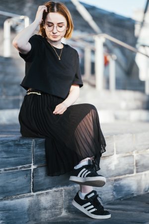 girl wearing a black t shirt, black pleated skirt, trainers and gold bee necklace with short ginger hair and round glasses