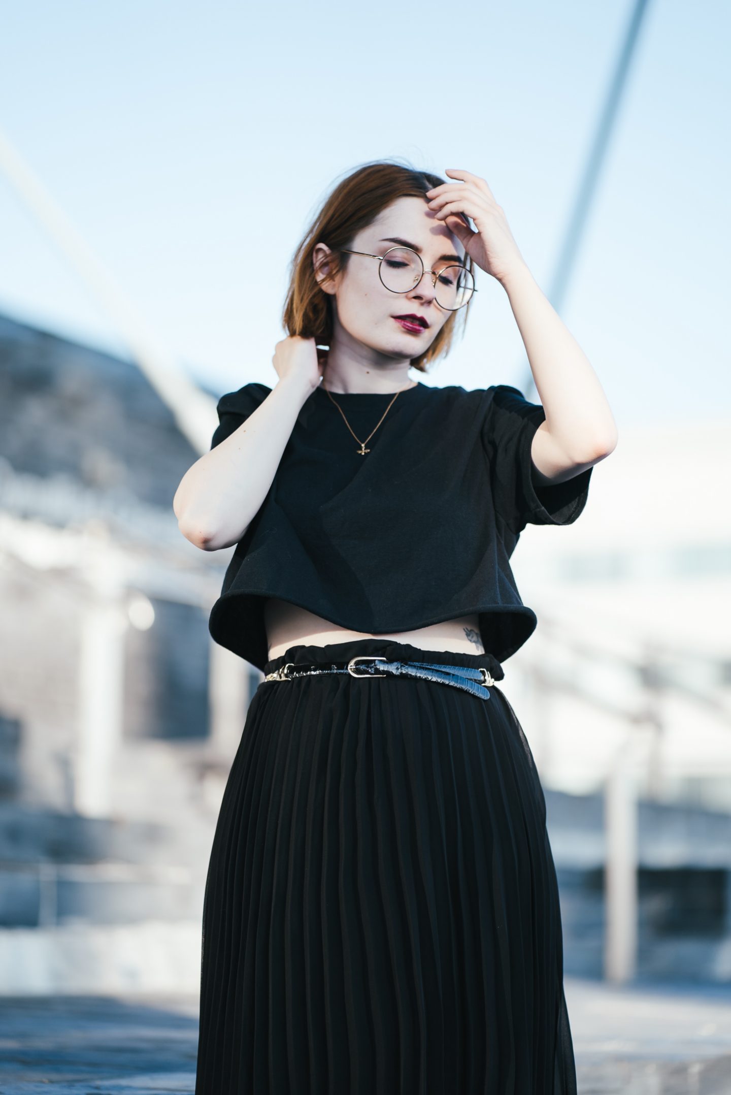 girl wearing a black t shirt, black pleated skirt and gold bee necklace with short ginger hair and round glasses