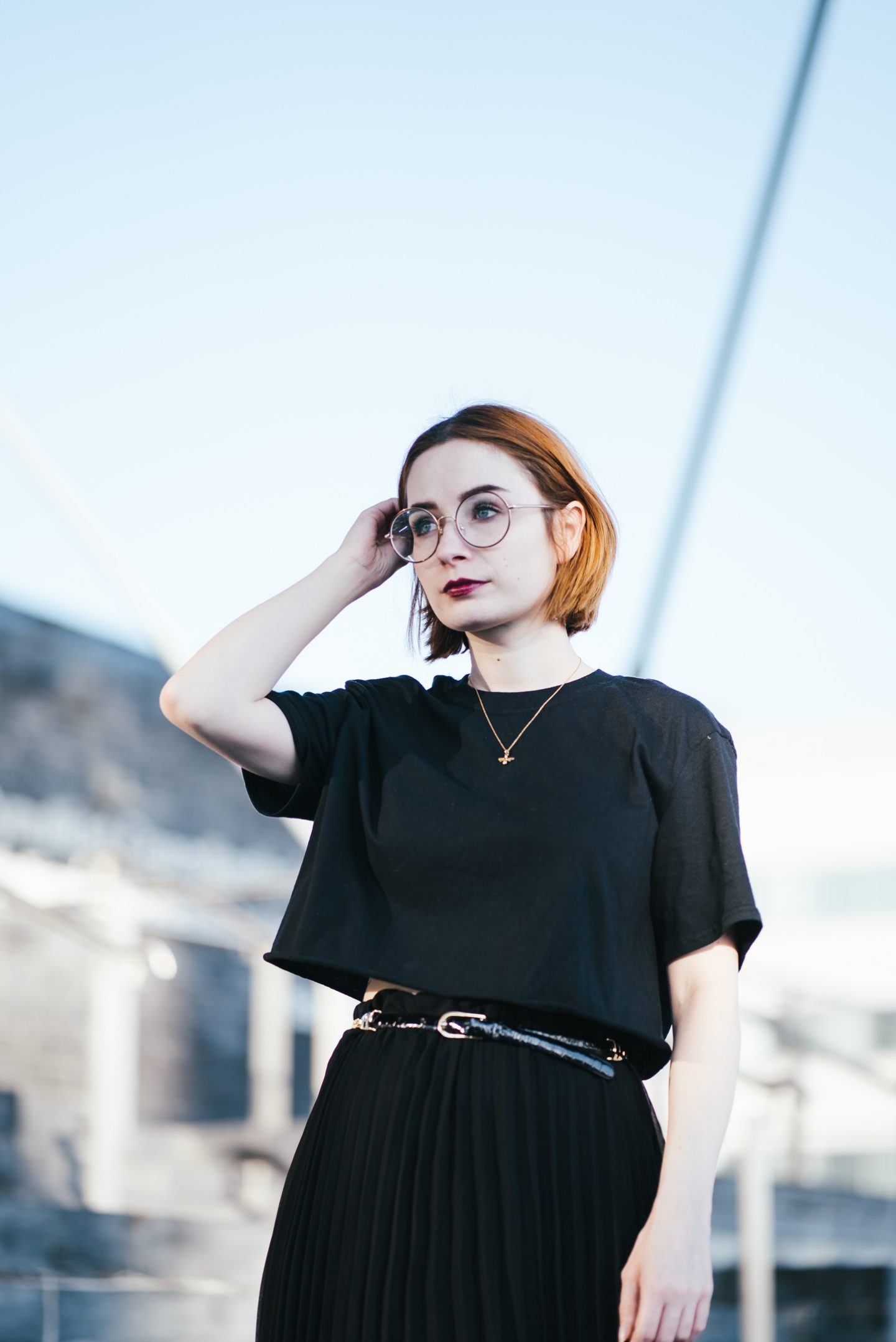 girl wearing a black t shirt, black pleated skirt and gold bee necklace with short ginger hair and round glasses
