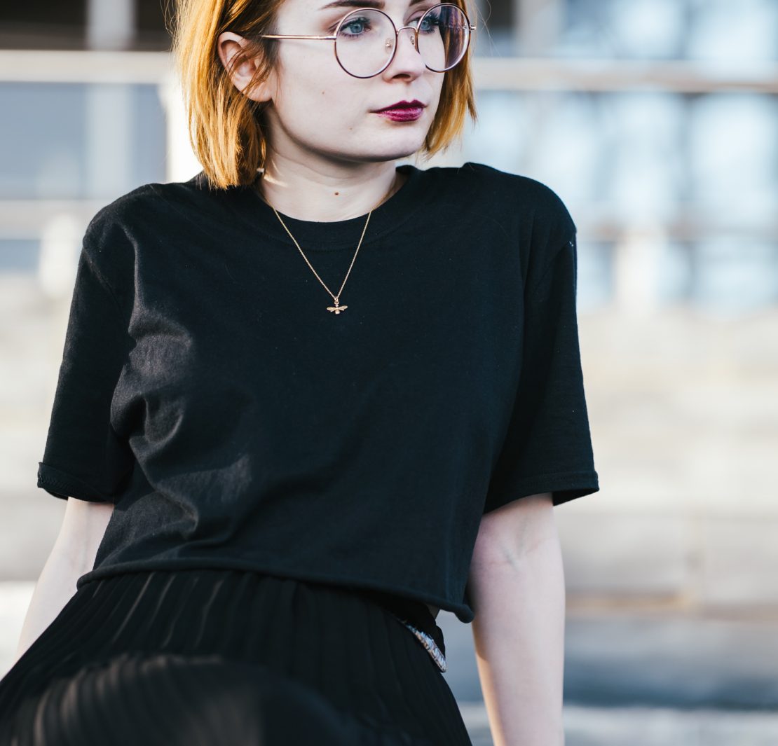 girl sitting down wearing a black t shirt and gold bee necklace with short ginger hair and round glasses