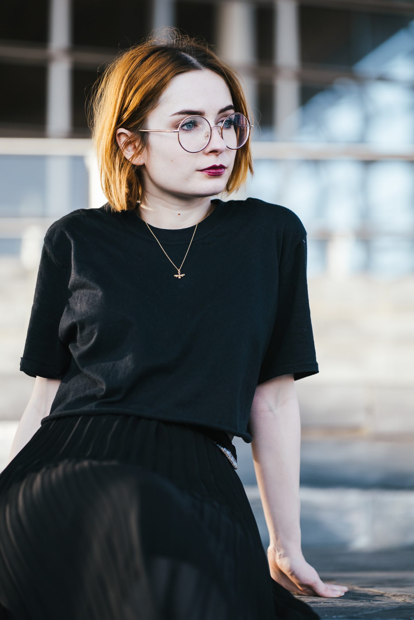 girl sitting down wearing a black t shirt and gold bee necklace with short ginger hair and round glasses