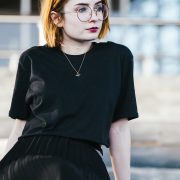 girl sitting down wearing a black t shirt and gold bee necklace with short ginger hair and round glasses