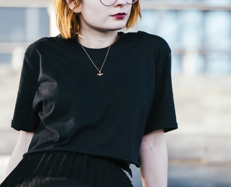 girl sitting down wearing a black t shirt and gold bee necklace with short ginger hair and round glasses