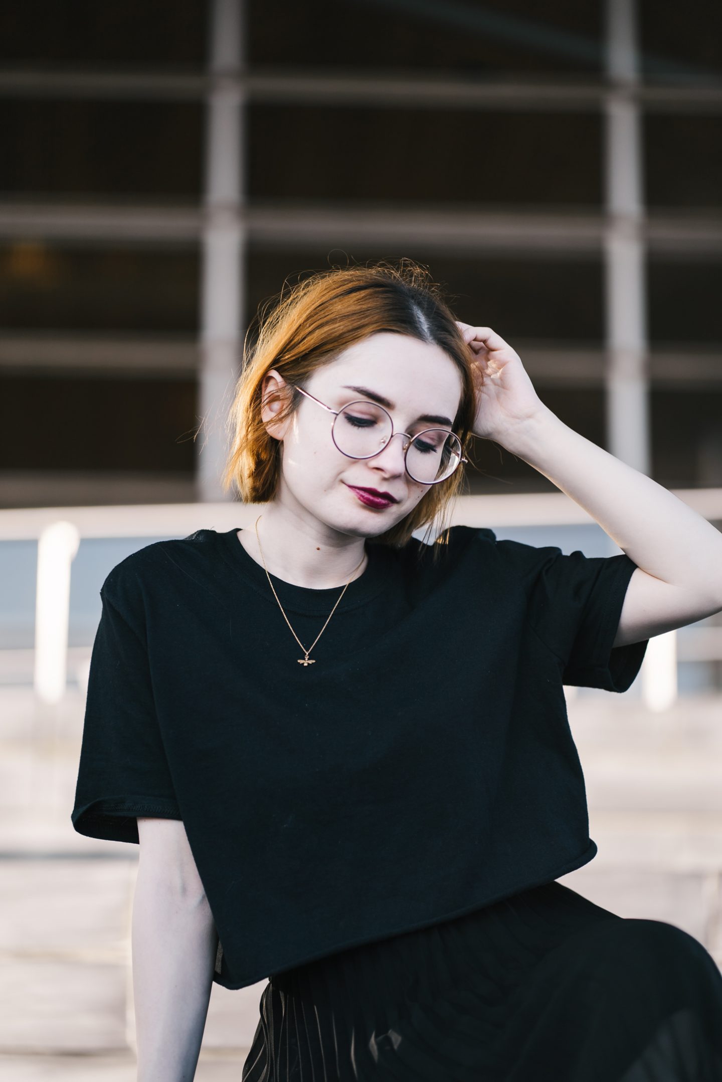 girl wearing a black t shirt, black pleated skirt and gold bee necklace with short ginger hair and round glasses