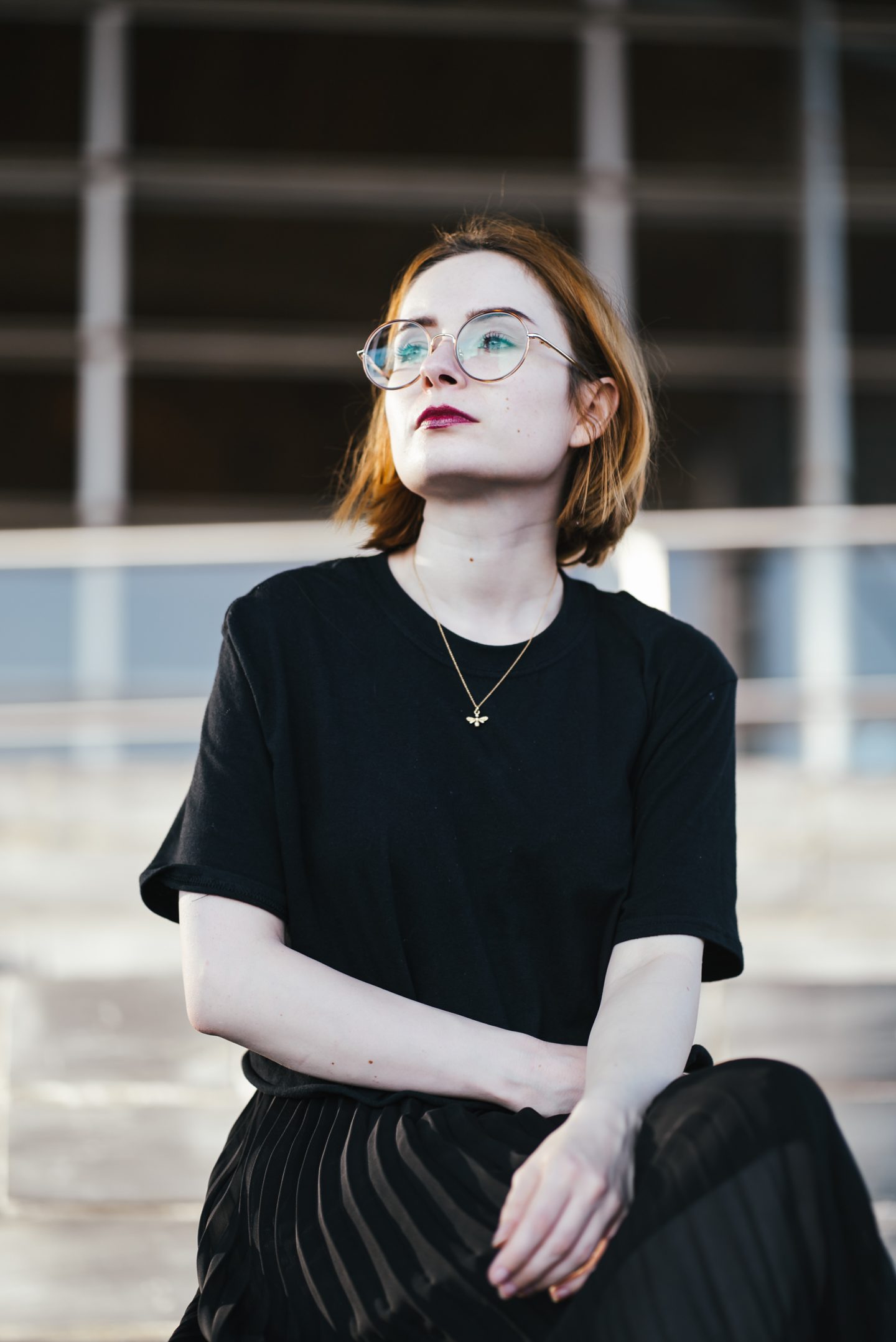 girl wearing a black t shirt, black pleated skirt and gold bee necklace with short ginger hair and round glasses