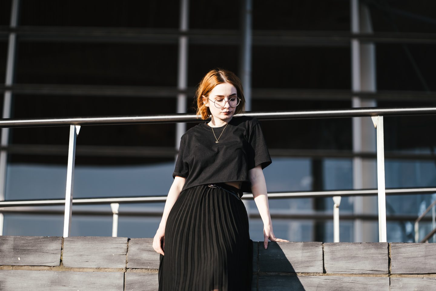 girl wearing a black t shirt, black pleated skirt and gold bee necklace with short ginger hair and round glasses