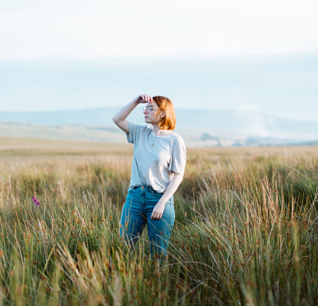 girl stood in field of long grass at sunset with hand to face to shield the sun. Planning the best staycation for a cheap UK holiday