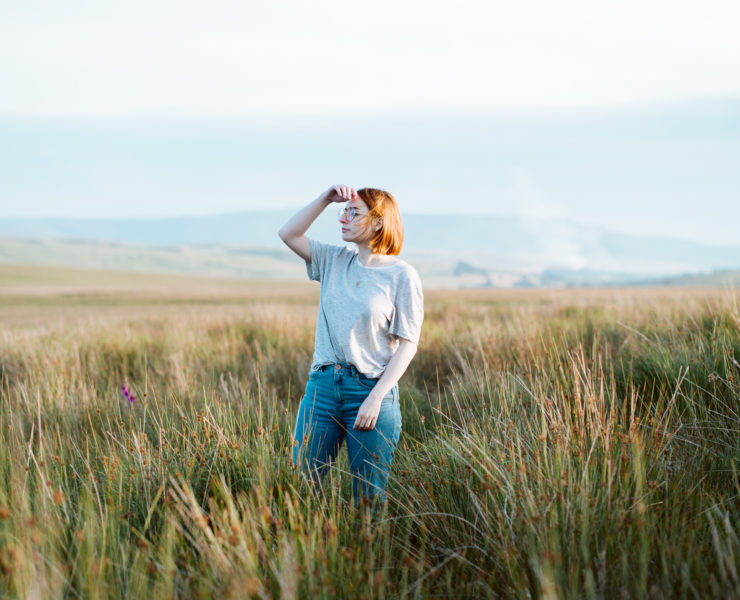 girl stood in field of long grass at sunset with hand to face to shield the sun. Planning the best staycation for a cheap UK holiday