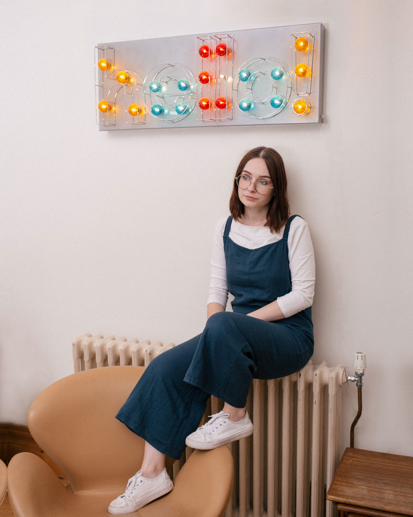 girl sat on radiator with feet resting on a chair below a light up sign that says hello
