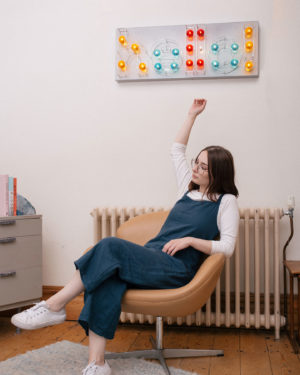 girl slouching in a 70s style chair below a neon sign that says hello