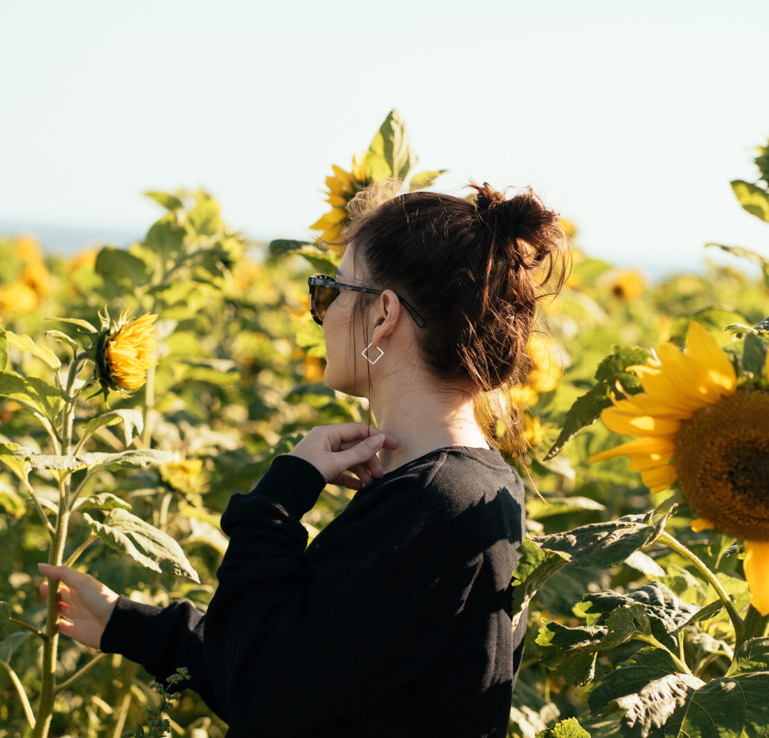 girl standing in a sunflower field at sunset wearing sunglasses, a black jumper and black shorts. Tips for a sustainable wardrobe