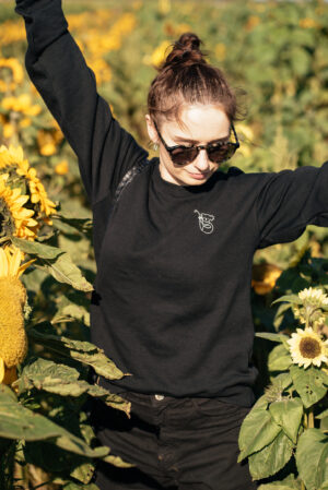 girl standing with arms up in the air in a sunflower field at sunset wearing sunglasses, a black jumper and black shorts. Tips for a sustainable wardrobe